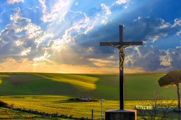 Jesus Christ on the cross by the road in the fields at sunset, with drammatic clouds and sun rays...