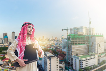 Obraz na płótnie Canvas Portrait of young arabian businessman holding the briefcase and standing in downtown with commitment to success with building background