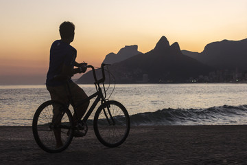 Man on a bicycle watching the sunset behind Dois Irmãos rock on Ipanema beach