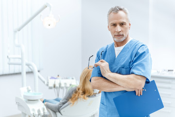 senior dentist in uniform with clipboard in dental clinic with patient behind