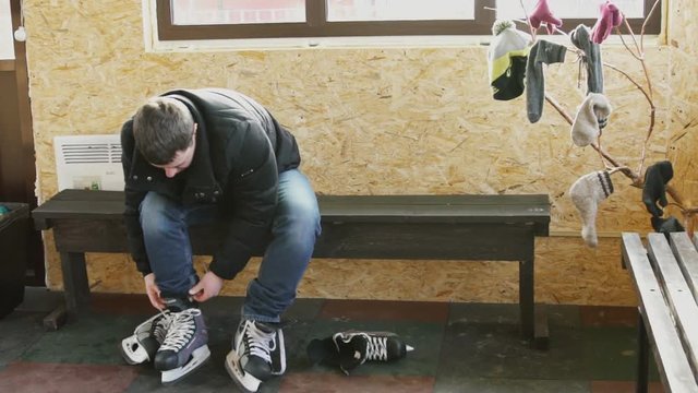 man dresses hockey skates for ice skating in locker room. male with dark hair in jeans, black jacket ties up long shoelaces. Preparation of footwear for winter entertainment. Slow motion of walker