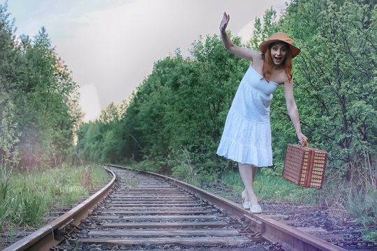 girl in a white sundress and wicker suitcase walking on rails