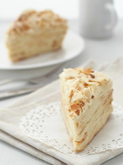 Piece of multi layered cake close-up. Mille feuille dessert. Crumbs decorated torte on white doily upon wooden table. Food background, blur. Selective focus on the front.