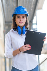 Vertical portrait of young beautiful architect with blue hardhat, protective headphones around her neck and clipboard among scaffolding. Selective focus