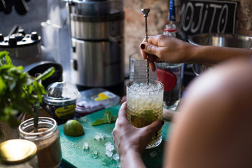 Young female bartender mixologist creates a fresh mojito out of lemons limes and crushed ice in a trendy bar full of character