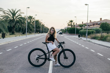 Fototapeta na wymiar Young female professional cyclist mountain biker seated in middle of road while sun is setting wearing beautiful white skirt smiling at camera