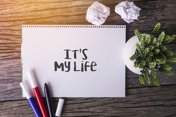 It's My Life word with Notepad and green plant on wooden background.