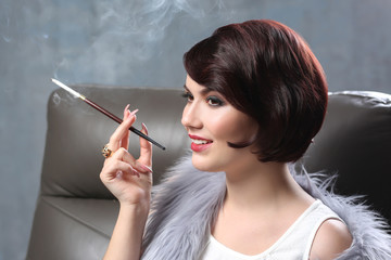 Young woman sitting in armchair and smoking with cigarette holder on color background