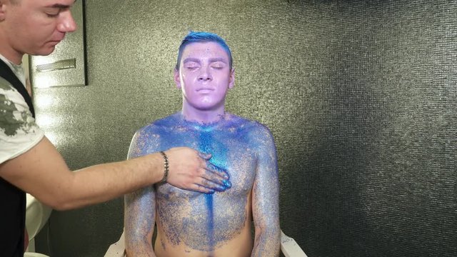 Body art artist covering the young man's body with blue sparkles