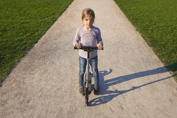 Beautiful child blond girl driving scooter on rural road outdoor in park