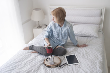Girl with cup in her hand, kettle and tablet on a white bed looking in window