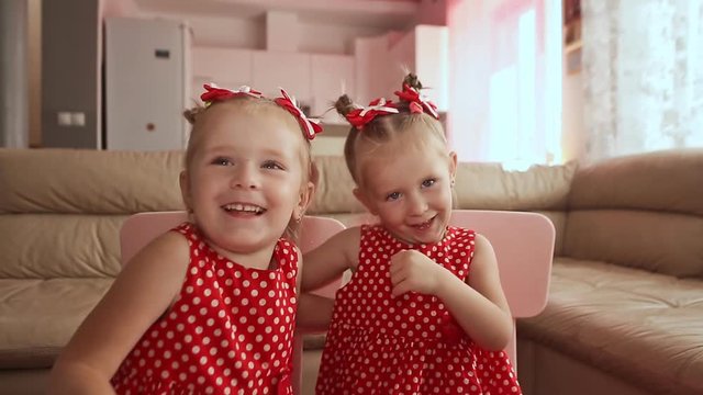 Two cute twin sisters are dressed in red polka-dot dresses. Playing together kiss each other smiling.