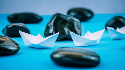 Two White Paper Ships in Sigle File between Abstract Black Rock Stones on Blue Background. Side Shot