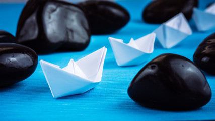 Leadership lead further white paper ships between stones on blue background. Right to left moving. low Angle of Shot. Blue Cold Colourtone