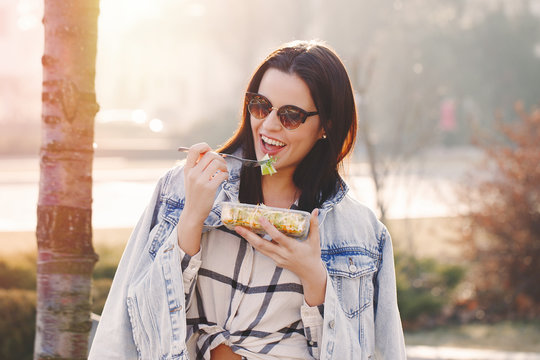 Young brunette woman in sunglasses eating fresh salad outdoor
