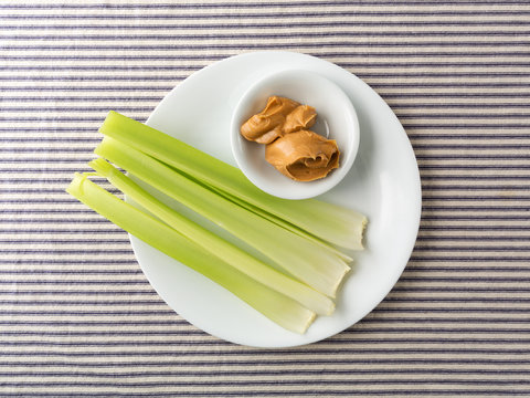 Top view of celery stalks with peanut butter in a small bowl on a white plate atop a blue striped tablecloth.
