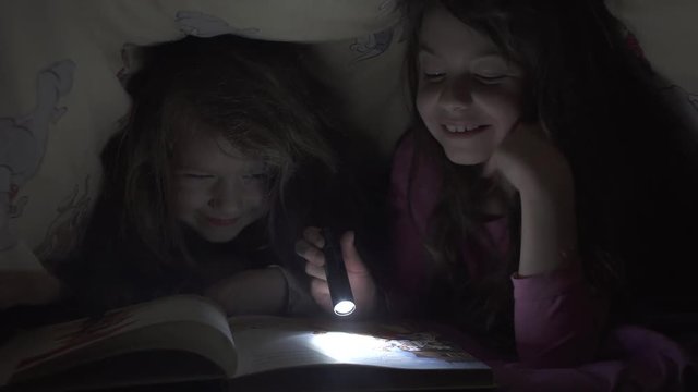 Children read a book under a blanket at night. Two little sisters hid under the bungle and do not want to sleep