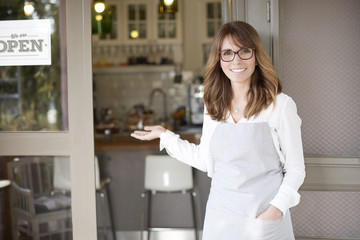 Proud small coffee shop owner businesswoman. Shot of  a happy barista woman standing at doorway and inviting to her coffee shop. Small business. 