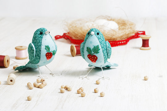 Closeup a two handmade birds sewed from a cloth stands on a white wooden table surrounded by handicraft materials. Shallow focus and blurred background.