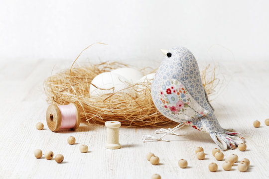 Closeup a handmade bird sewed from a cloth and nest on a white wooden table surrounded by handicraft materials. Shallow focus and blurred background.