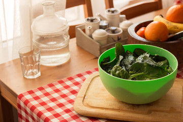 Spinach leaves in green plate on wooden chopping board in kitchen with oranges and glass water decanter in frame