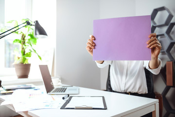 Young businesswoman hiding head behind paper at workplace in office.