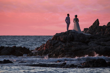 Just married beach couple at sunset with bride and groom standing close and beautiful pink sunset over the ocean.  Waves and rocks, with room for copyspace.  Romance and true love.