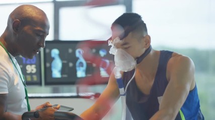  Man with breathing equipment on exercise bike having fitness levels analyzed by sports trainer.  - Powered by Adobe