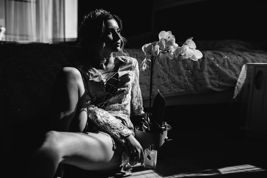 Beautiful woman in lingerie showing long legs sexy pose leaning at bed over her bedroom with big window, flowers. Sexual memories. Bride boudoir. Wedding morning.