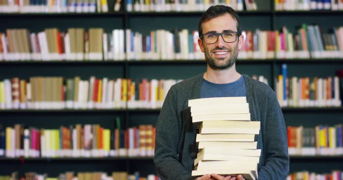 Portrait of a beautiful young man smiling happy in a library holding books after doing a search and after studying. Concept: educational, portrait, library, and studious.