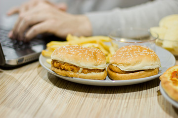A man works at a computer and eats fast food. unhealthy food: Burger, sauce, potatoes, donuts,chips.