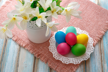 Obraz na płótnie Canvas A white dish with bright painted Easter eggs. Artificial white flowers in the white vase. All worth it on a napkin on wooden background.