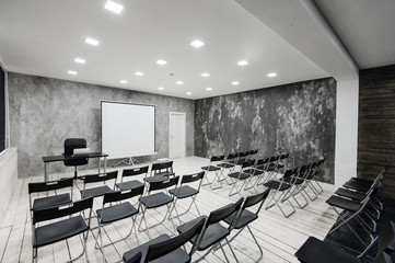 Room for lecture with a lot of dark chairs. Walls are white, loft interior. On the right there is a door. On the background there is a table with a laptop.