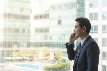 Mature and confident business executive looking looking out of large windows at a view of the city below, from the top floor of an office building, while talking on his mobile phone