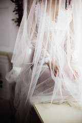 beautiful girl in white transparent boudoir gown of tulle and lace closed with a veil sitting on the windowsill near the window