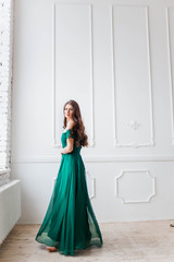 beautiful girl in a long emerald green dress standing near the white background in the Studio window looks into the camera barefoot