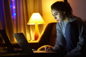 woman watching laptop at winter evening in dark room with window as background. illuminated garland in the window
