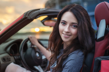 Pretty young brunette woman driving luxury red cabriolet car at the sunset. - obrazy, fototapety, plakaty