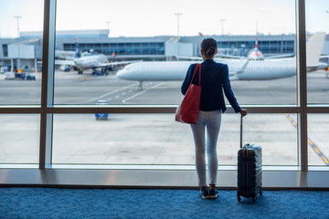 Businesswoman traveling in airport. Woman looking through the window at tarmac and planes waiting...