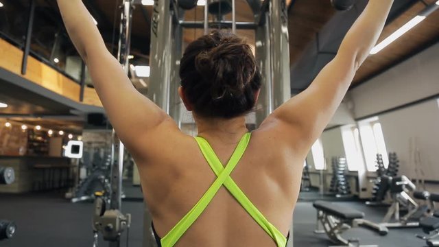 Young woman do exercise using training machine in gym. View from back. Video focus on shoulders and arms of sportswoman, no face, only head with red hair tied in bun not to interfere. Her sportswear