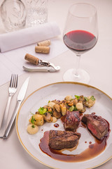 A plate with beef fillet served on a table with a white cloth. A garnish from mushrooms and hazelnuts with a celery. A glass of red wine with tableware.