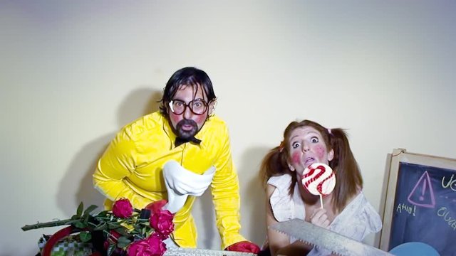 A weird salesman hiding his face under a white mask, together with his female assistant licking a lollipop. Funny comedy foolish characters.
