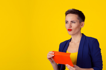 Thoughtful woman holding red envelope with money cash in it thinking what to do next, daydreaming planning how to spend them isolated yellow background wall