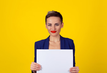Fototapeta premium Smiling woman holding the piece of paper isolated yellow background, blue shirt red lips short hair. Head sot, portrait, horizontal image studio shot one model