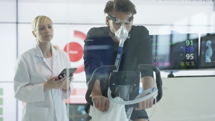  Man with breathing equipment on exercise bike having fitness levels analyzed by sports scientist in white coat. 