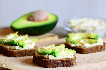 Hummus avocado sandwiches on a wooden board, avocado half, hummus in a glass bowl. Vegetarian sandwiches cooked with rye bread, avocado slices, hummus and roasted sesame seeds