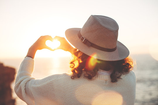 Young traveling female in hat enjoing the adventure, woman hipster making heart symbol with hands at sunset. Intentional lens flares