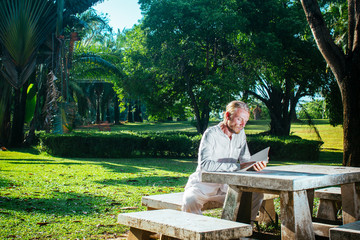 Male reading book at table in park. Young man in white clothing sitting at stone table in tropic park and reading book.