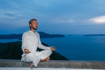 Male in lotus pose . Man in white clothing meditating in lotus pose sitting on fence in dusk with sea on background.