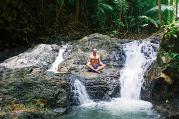 Man practicing yoga among waterfall. Young male sitting in lotus pose on rock with waterfall streams  near in tropical forest.